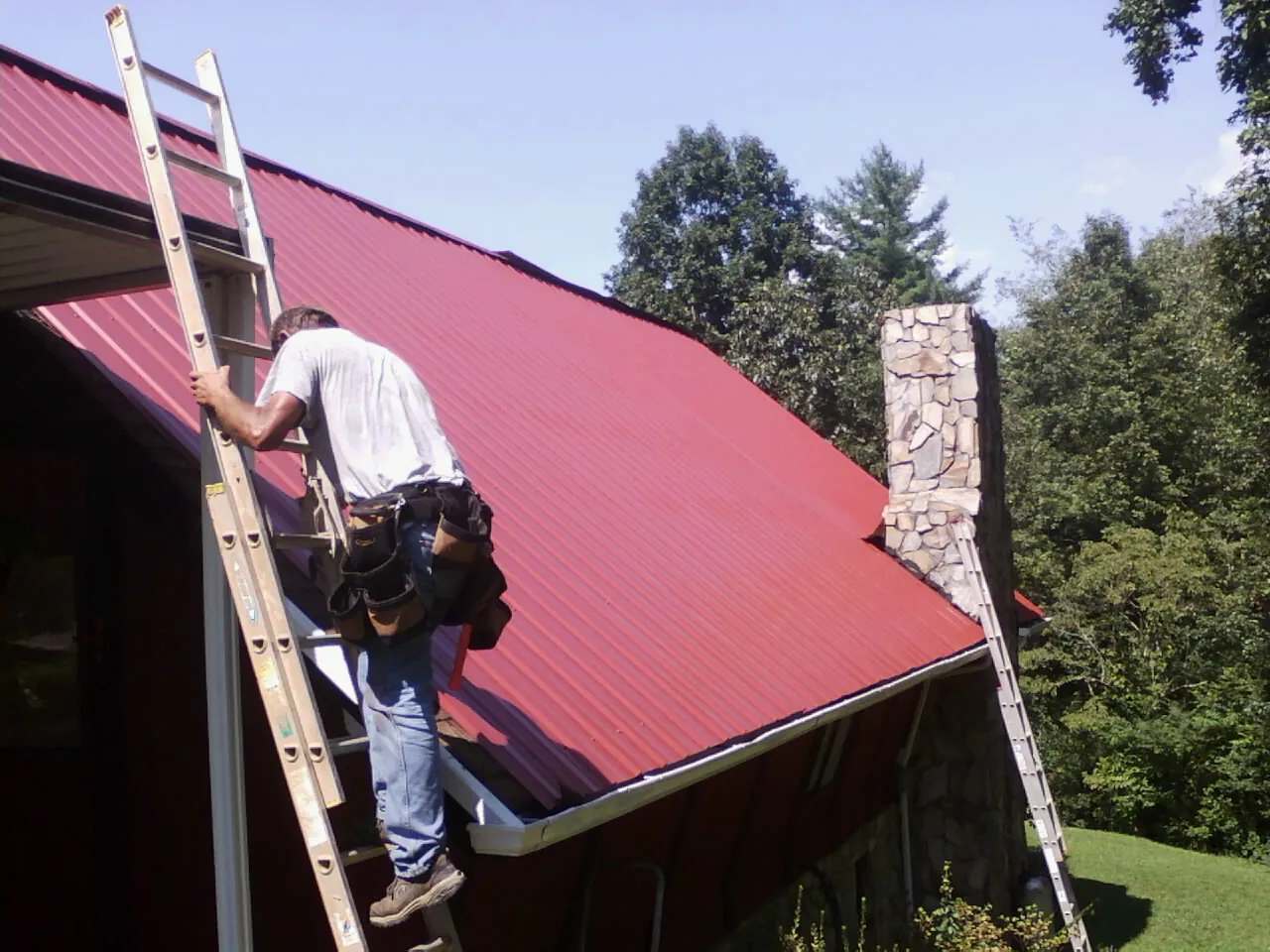 Roofer on a ladder beside a red metal roof and stone chimney — Soffit & Fascia Repair by Roofing Brothers in Sharon Center
